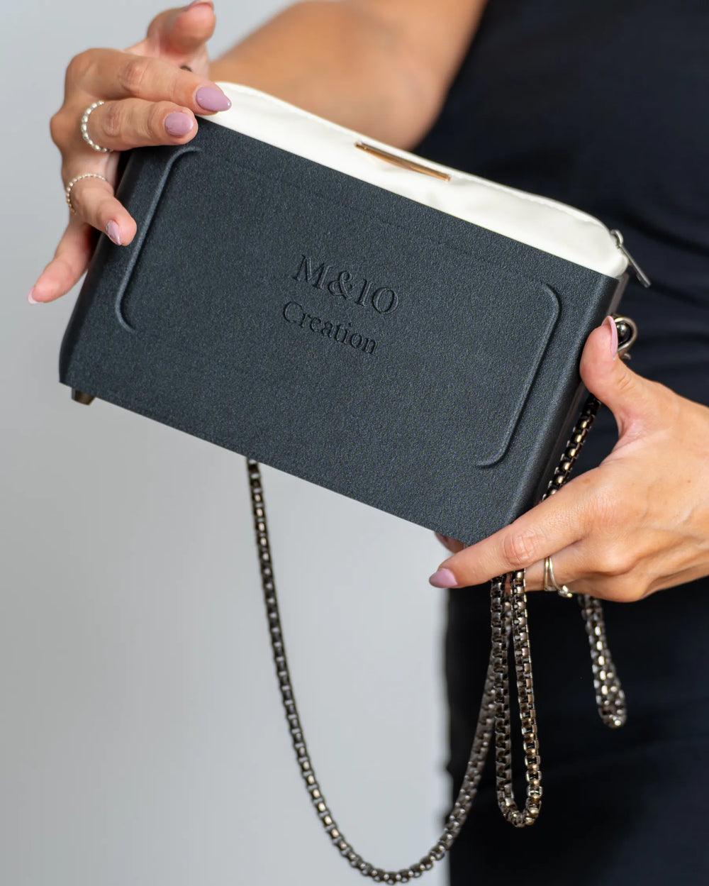 Two hands hold the Black Stone bag at an angle, M&IO Creation raised on the front; the cream pouch shows above the edge, the chain hanging below.