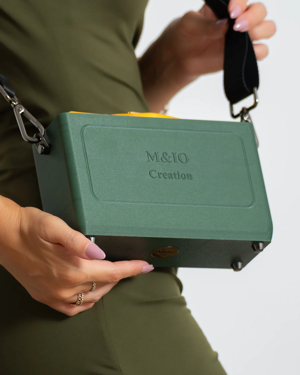 Close up, the Ulivi di Puglia bag held in front of the hip, the black strap pulled towards the shoulder: M&IO Creation raised on the front, the yellow pouch showing above the edge.
