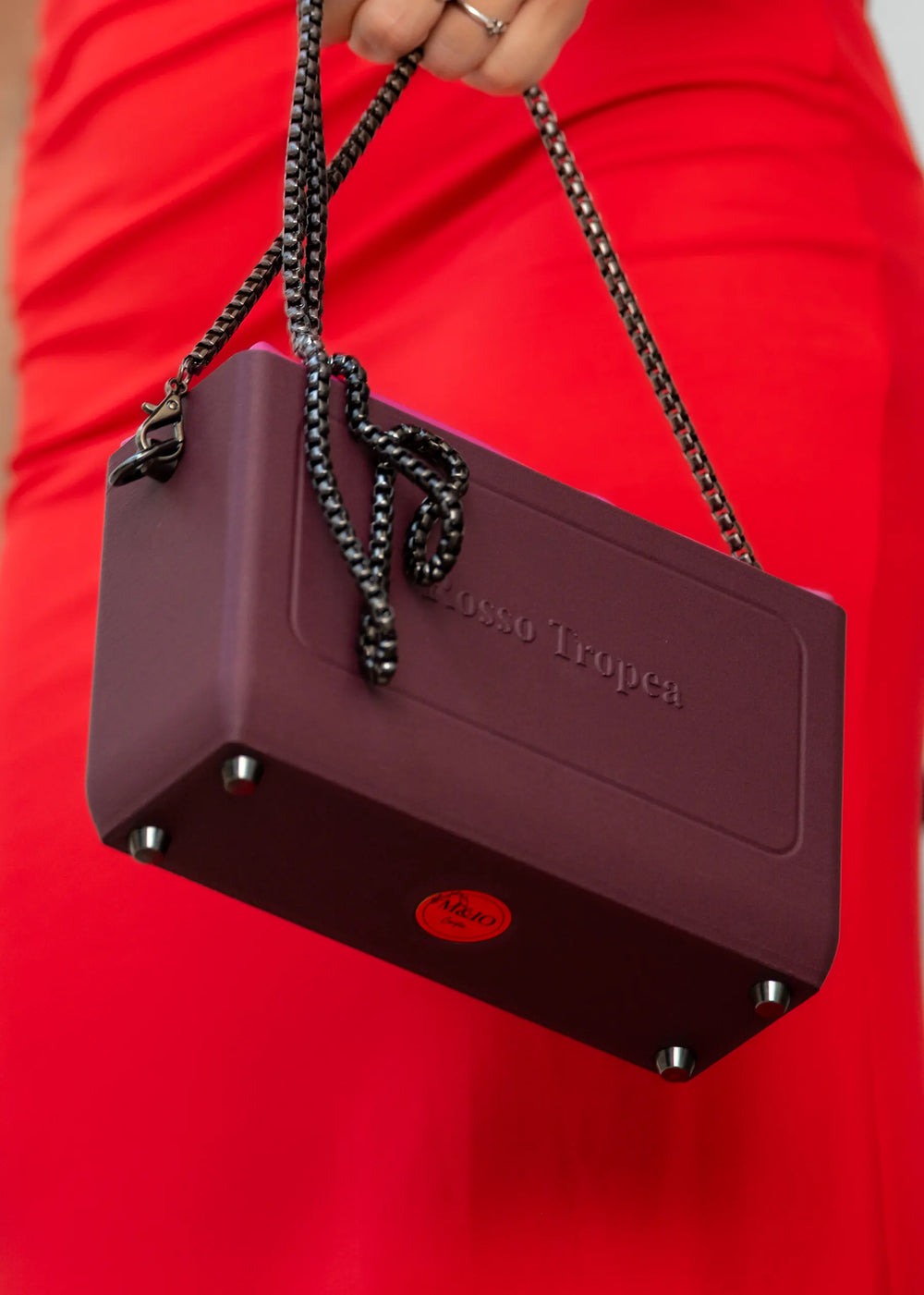 The Rosso Tropea bag hanging from its chain, held in front of a red dress: the raised lettering on the front, the medallion underneath.